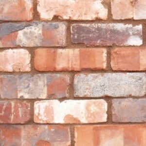 A panel of reclaimed brick slips. The blend of bricks has alarge variation in colour and texture. There are light orange, mid-red and plum coloured bricks with an occasional white. The bricks have old mortar on the faces and the edges of the bricks are chipped and cracked. The panel is pointed in a mid brown pointing mortar